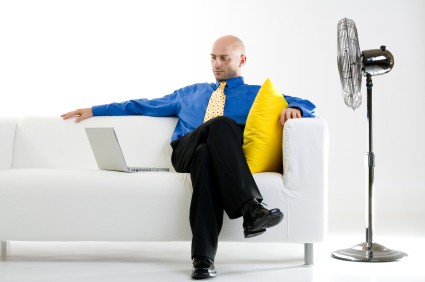 Businessman relaxing on a white couch with a laptop computer beside him to his right. A large standing fan is to his left, pointed in his direction. Isolated on a white background.
